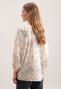 Cremefarvet bluse med sorte blomster- og paisleymønstre, bløde ærmer og samling ved halsudskæringen. Båret med mørkegrå bukser.