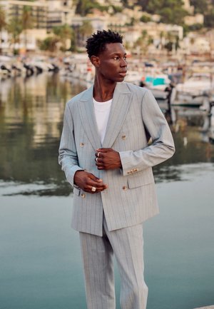 Mann in hellgrauem Nadelstreifenanzug und weißem Hemd steht nahe am Wasser mit Booten und Gebäuden im Hintergrund, schaut zur Seite.