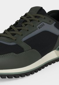 Gros plan d'une chaussure de sport vert foncé et noire avec un tissu mesh texturé, des lacets verts et le marquage "BOSS" sur le panneau latéral.