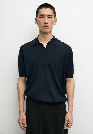 Homme aux cheveux noirs courts portant un polo bleu marine à manches courtes et un pantalon noir, debout devant un fond clair uni.
