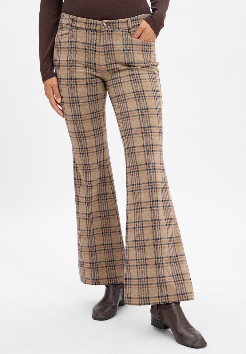 Pantalons évasés à carreaux beige avec des lignes bleu foncé ; dotés de poches avant et d'une fermeture à bouton ; portés avec des bottines marron.