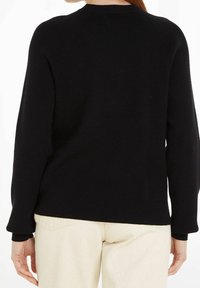 Person iført en sort langærmet sweater og off-white bukser, set bagfra mod en ensfarvet baggrund.