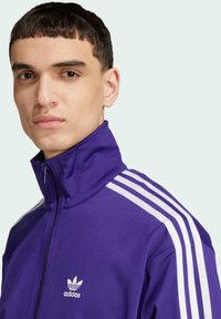 Lilla zip-up jakke med hvite striper på skuldrene, laget av glatt stoff. Har høy krage og et Adidas-logo på brystet.