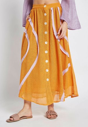 Narančasta pletena midi suknja s bijelim gumbima, s ružičastim vrpcama kao akcentima. Uparena s smeškim naboranim kliznim sandalama.