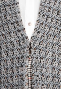 Grå stickad cardigan med ett strukturerat mönster, prydd med orangea och svarta prickar. Sidodragkedja med ett rutigt tygaccent synligt under.