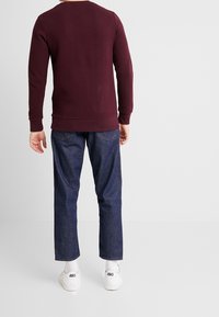 Persona in piedi di spalle che indossa un maglione testurizzato color bordeaux, jeans blu scuro, calzini bianchi e sneakers bianche ASICS, su uno sfondo neutro.