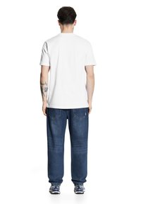 Biała bawełniana koszulka z krótkim rękawem, połączona z luźnymi niebieskimi dżinsami. Widoczne tatuaże na lewym ramieniu, zestawione z trampkami.