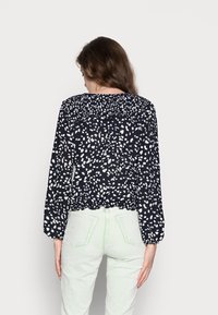 Schwarze und weiße gemusterte Bluse mit langen Ärmeln und gerafftem Ausschnitt, getragen mit hellgrünen Cordhosen, die ein kurzes Design zeigen.