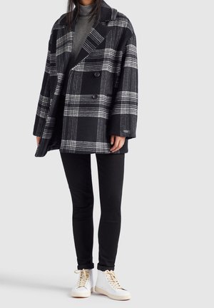 Person iført en sort og hvid ternet oversized frakke, grå højhalset trøje, sorte smalle bukser og hvide høje sneakers.