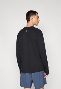 Chemise noire à manches longues avec texture côtelée, dotée d'un col rond et d'ouvertures sur les épaules. Portée avec un short bleu.