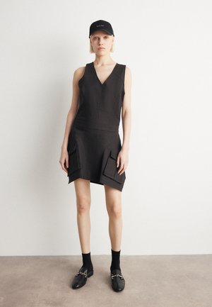 Svart ärmlös klänning med V-ringning, med en figurnära bodice och utsvängd kjol. Två stora fickor på kjolen och en texturerad yta. Svarta loafers.