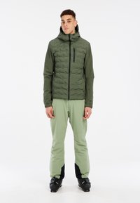 Veste d'isolation verte avec capuche, présentant un design matelassé et une poche zippée sur la poitrine, associée à un pantalon vert clair et des bottes de ski noires.