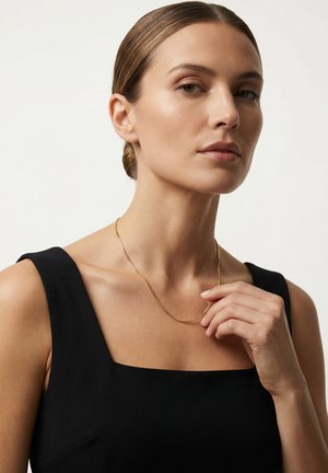 Femme aux cheveux tirés en arrière, portant un haut noir sans manches, tenant près de sa clavicule un collier fin en chaîne dorée.