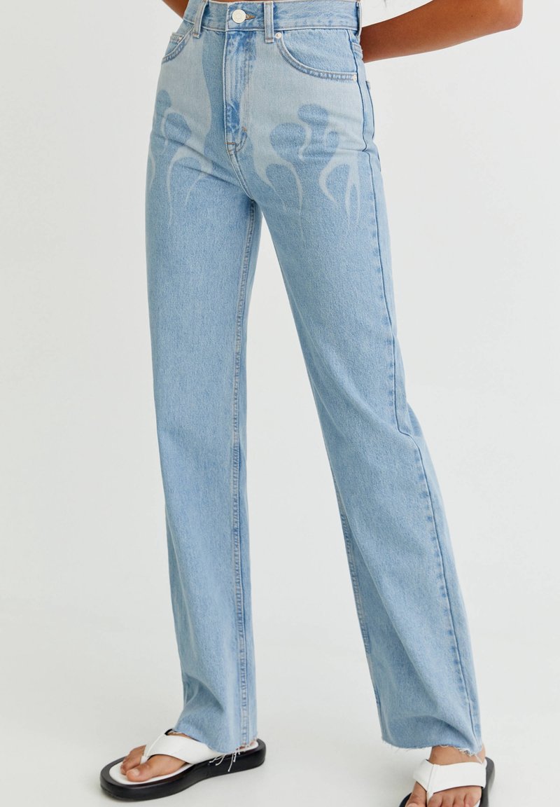 Personne portant un jean bleu clair taille haute à motifs abstraits délavés et des sandales blanches, debout sur un fond uni.