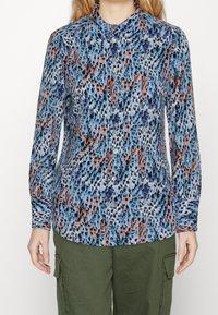 Donna che indossa una camicia a maniche lunghe con bottoni, caratterizzata da un motivo astratto blu, nero, bianco e arancione, abbinata a pantaloni cargo verde oliva.