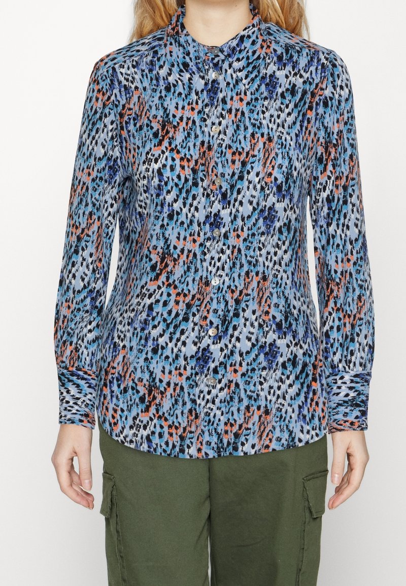 Donna che indossa una camicia a maniche lunghe con bottoni, caratterizzata da un motivo astratto blu, nero, bianco e arancione, abbinata a pantaloni cargo verde oliva.