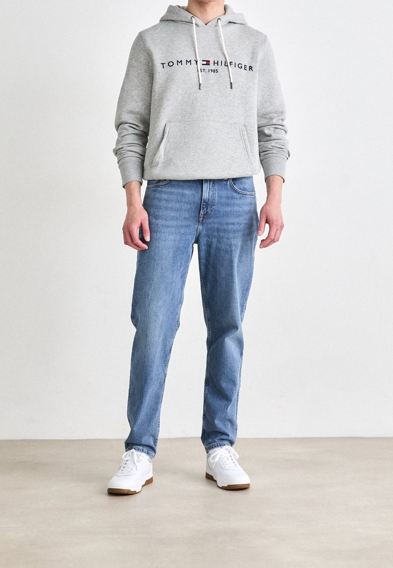 Felpa grigia in cotone con cappuccio e cordino, che presenta il logo "TOMMY HILFIGER". Indossata con jeans a gamba dritta azzurri e sneakers bianche.