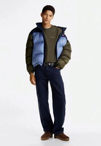 Young man standing with hand in pocket, wearing blue and green puffer jacket, olive green shirt, dark pants, and brown shoes against white background.