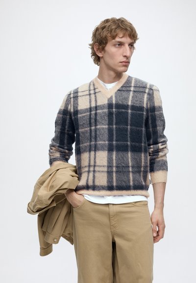 Jeune homme portant un pull à carreaux beige et gris foncé tenant une veste couleur sable dans une main, avec un pantalon beige et une chemise blanche en dessous.