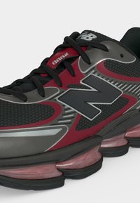 Μαύρο και κόκκινο αθλητικό παπούτσι New Balance με ορατή ενδιάμεση σόλα απορρόφησης κραδασμών και μαύρα κορδόνια, που δείχνει το λογότυπο της μάρκας στο πλάι και στη γλώσσα.