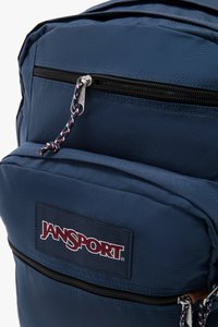 Marineblå Jansport ryggsekk med slitesterkt stoff, to glidelåslommer og en flettet trekkstang med merkevarelogo.
