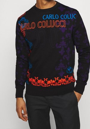 Homme portant un sweat-shirt noir avec des motifs floraux rouges, bleus et violets et le texte "CARLO COLUCCI" en bleu et rouge.