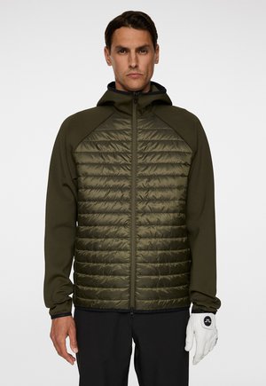 Man in een olijfgroene gewatteerde jas met capuchon en zwarte broek, rechtop staand met één witte handschoen aan de rechterhand.