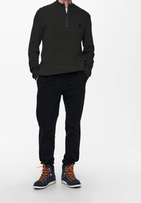 Dunkelgrüner, gerippter Pullover mit halbem Reißverschluss, kombiniert mit schwarzen Hosen und marineblauen Stiefeln mit orangen Schnürsenkeln und weißen Sohlen.