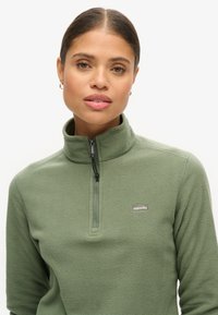 Groene fleece pullover met hoge kraag, ritsluiting en logo op de borst. Zachte textuur en ontspannen pasvorm, geschikt voor lagen.