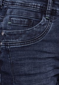 Donkerblauwe denim jeans met een voorzak, subtiele groene stiksels en klassieke vijf-pocket styling met metalen knoopsluiting.