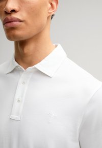Witte polo shirt met een klassieke kraag, knoopsluiting en subtiel geborduurd logo op de borst. Soepele katoenweefseltextuur.