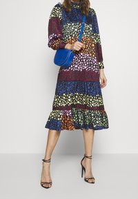 Värikäs, kuviollinen mekko pitkillä hihoilla, frillalla varustettu helma sekä sekoitus sinisiä, purppuran, vihreitä ja oransseja pisteitä; yhdistettynä siniseen olkalaukkuun.