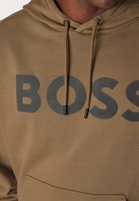 Felpa con cappuccio marrone con grande logo nero "BOSS", coulisse con punte metalliche e tasca a marsupio frontale. Tessuto morbido e strutturato.