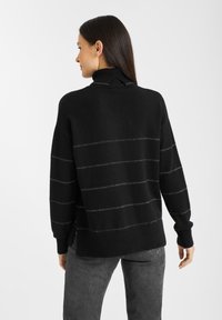 Vrouw in een zwart coltrui met dunne horizontale zilveren strepen, gecombineerd met donkergrijze jeans, met de rug naar de camera gekeerd tegen een eenvoudige witte achtergrond.
