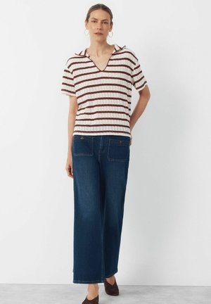 Kvinde stående iført en kortærmet hvid skjorte med brune striber og blå viddebukser, foran en ensartet lys baggrund.