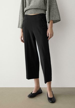 Woman wearing cropped black trousers with side button details, gray knit sweater, and black ballet flats standing on gray floor.