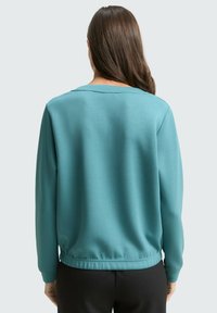 Morska zieleń bluzy z długimi rękawami, okrągłym dekoltem i elastycznym pasem. Gładka faktura, prosty fason, brak widocznych wzorów czy ozdób.