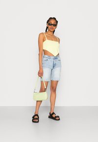 Top amarelo curto, calções de ganga, sandálias pretas e uma pequena bolsa verde. Apresenta alças finas, calções com a bainha desfiada e uma textura suave na bolsa.