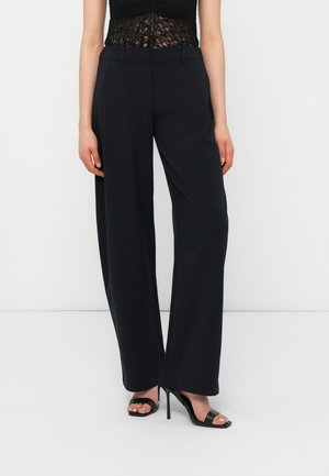 Γυναίκα που φοράει μαύρο ψηλόμεσο παντελόνι με φαρδιά πόδια και μαύρα σανδάλια με ψηλό τακούνι, στέκεται μπροστά σε λευκό φόντο.