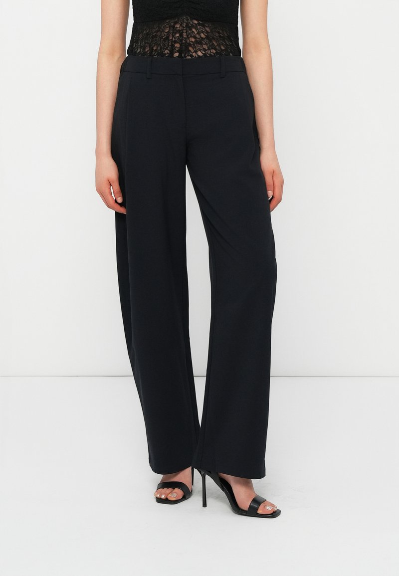 Femme portant un pantalon noir taille haute à jambes larges et des sandales à talons hauts noires, debout devant un fond blanc.