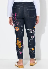 Vaqueros ajustados de mezclilla oscura con estampados gráficos coloridos que incluyen un tigre, un símbolo de paz y estrellas; presentan costuras en contraste y cinco bolsillos.