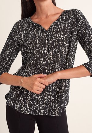 Blouse noire avec motif de points blancs dispersés, manches trois-quarts, col en V avec petit nœud, portée avec un pantalon noir slim et des bracelets fins.
