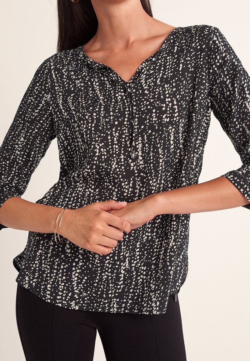 Blouse noire avec motif de points blancs dispersés, manches trois-quarts, col en V avec petit nœud, portée avec un pantalon noir slim et des bracelets fins.