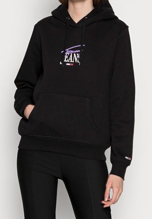 Osoba w czarnej bluzie z kapturem z przednią kieszenią i logo Tommy Jeans, w czarnych spodniach, stojąca na jednolitym tle.