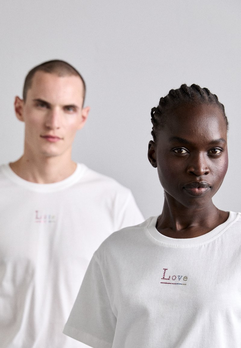 White cotton t-shirts with short sleeves, featuring colorful "Love" embroidery on the chest. Smooth texture, relaxed fit, minimal design.