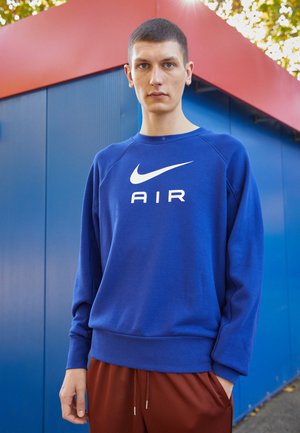 Niebieska bluza Nike z białym logo "AIR", z prążkowanym okrągłym dekoltem i mankietami. Nosi ją z brązowymi spodniami sportowymi. Tło jest niebiesko-czerwone.