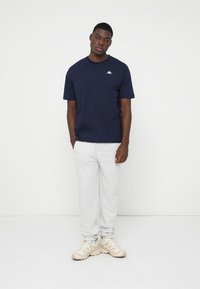 T-shirt de algodão azul-marinho com decote redondo e pequeno logótipo branco, combinado com calças de fato cinza-claro e ténis bege. Conjunto casual simples.