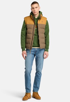 Gilet imbottito marrone e beige con zip frontale, abbinato a un cappuccio verde, jeans azzurri e stivali marroni con lacci.