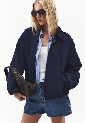 Blondynka nosi czarne okulary przeciwsłoneczne, granatową kurtkę, jasnoniebieską koszulę, biały tank top, jeansowe szorty i trzyma fakturowaną ciemnobrązową torebkę.