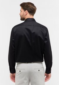 Zwarte longsleeve shirt met een gladde textuur, standaard kraag en knoopmanchetten, gezien vanuit de achterkant, gecombineerd met lichtgrijze broeken.